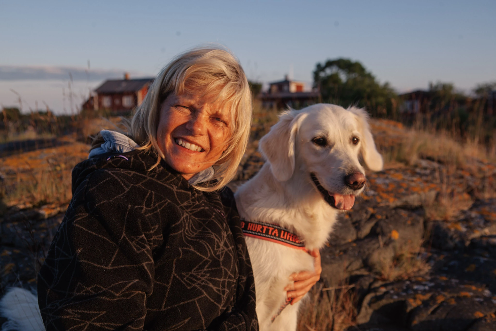 En äldre kvinna och en hund sitter tätt tillsammans i varmt kustljus.
