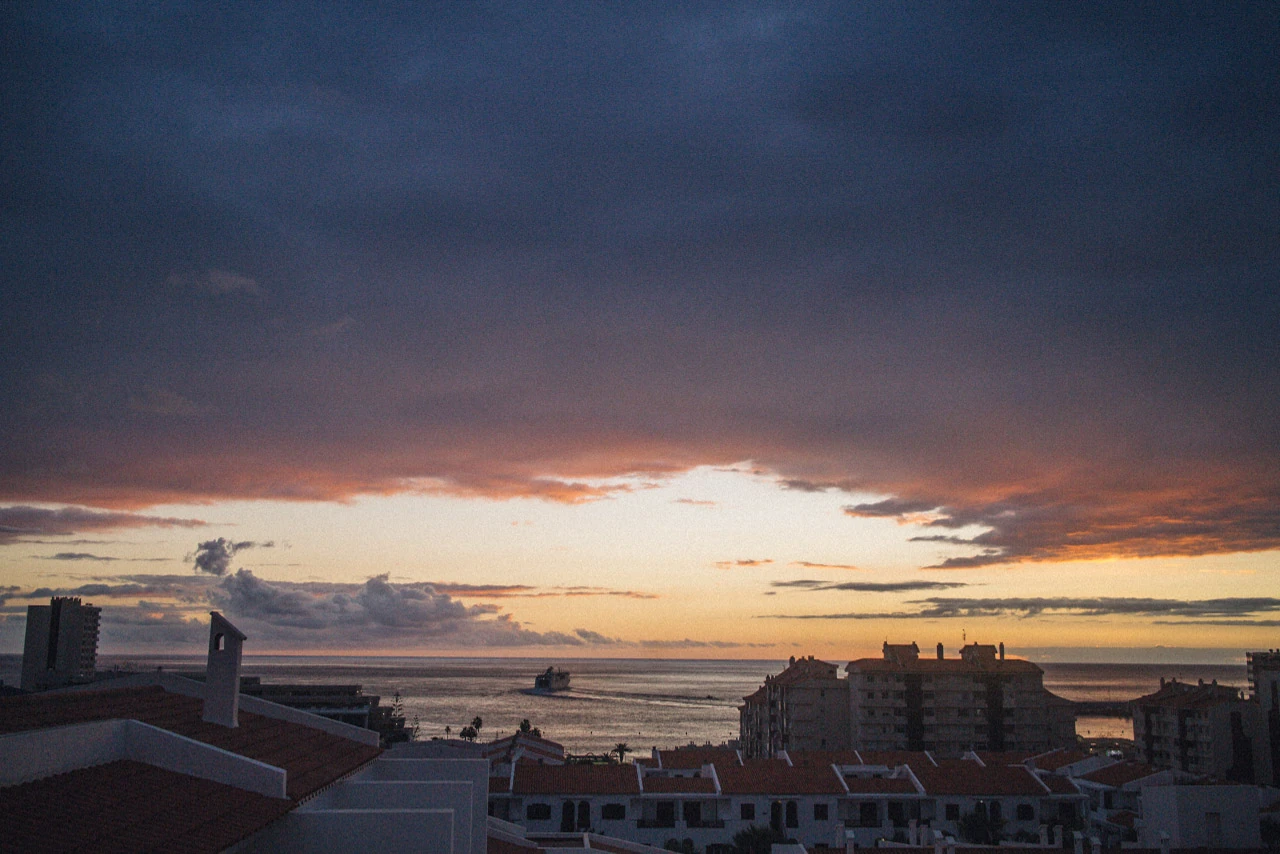 Dark rooftops in front of the sea and clouds in orange and blue twilight.
