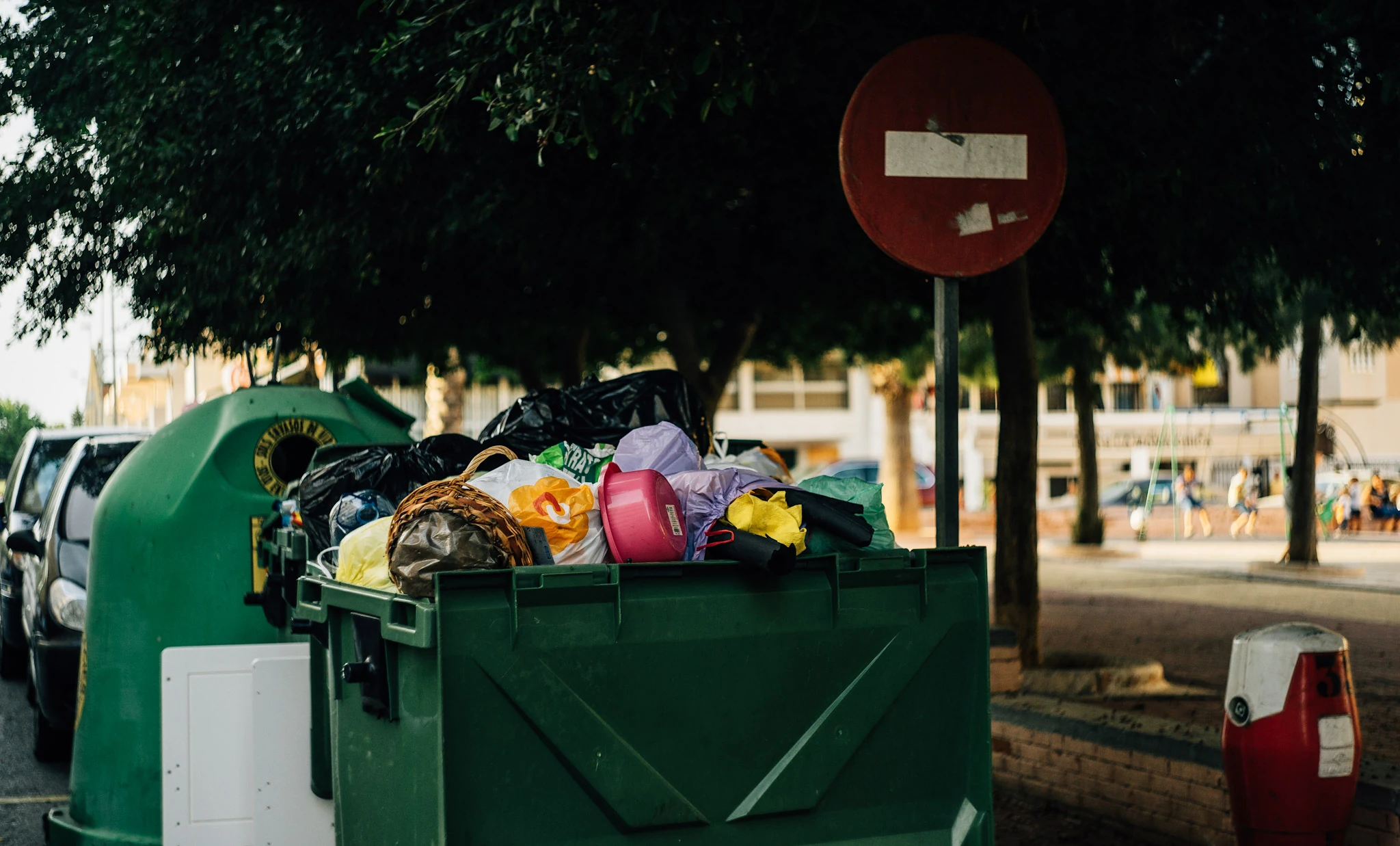Overflowing bins stand beneath a tree and a red traffic sign.