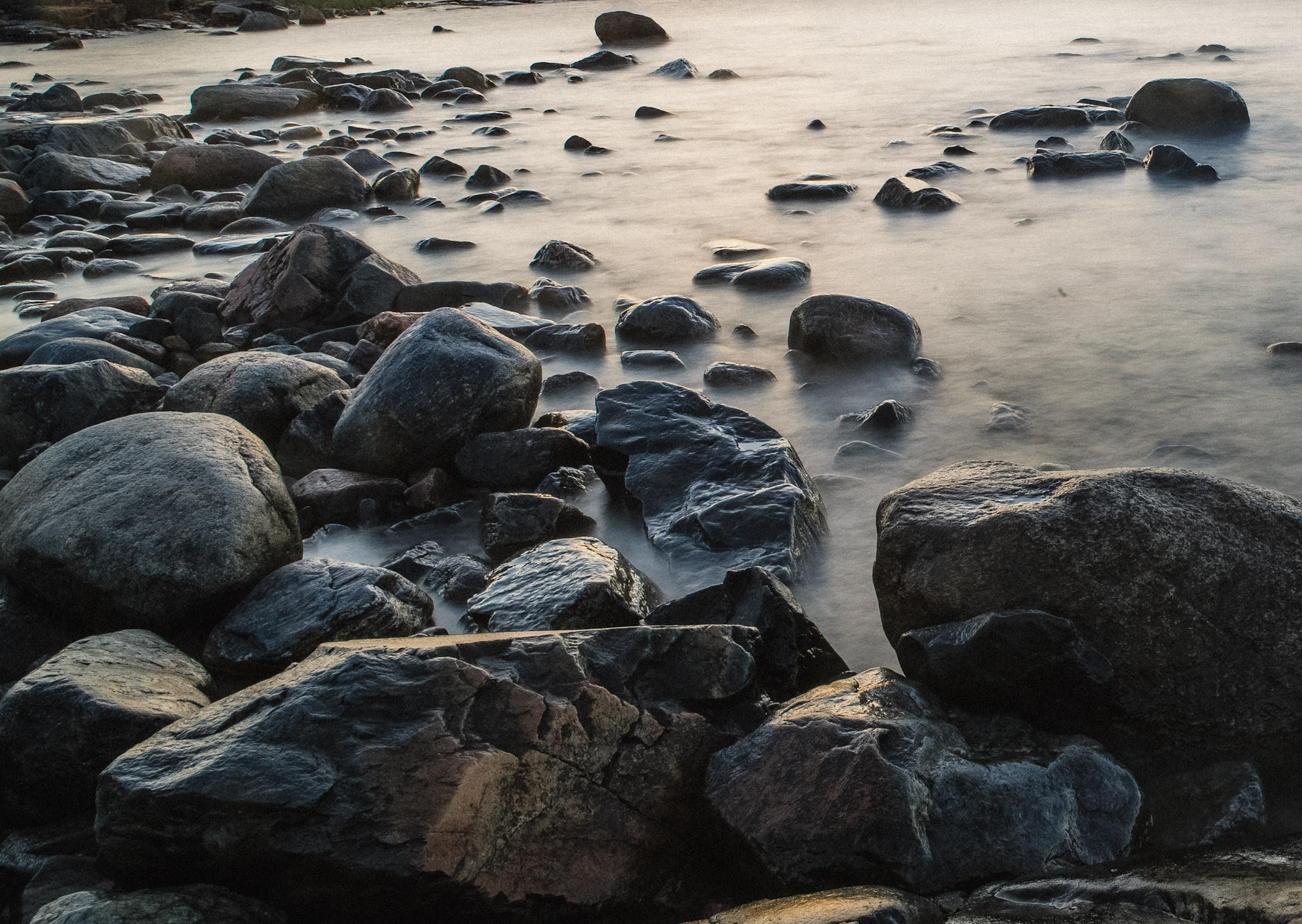 Mörka rundade stenar ligger i mjukt, ljust vatten vid en strand.