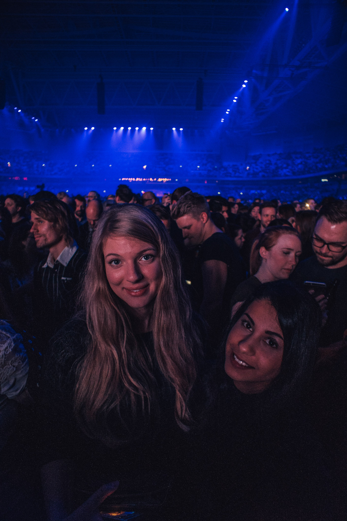 Two women in a concert crowd with blue stage lighting across the arena behind them.