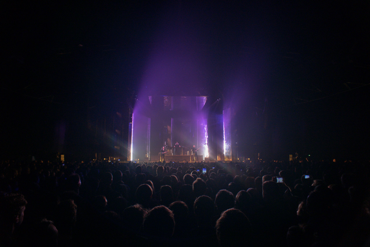A concert stage in violet light beyond a dense audience.