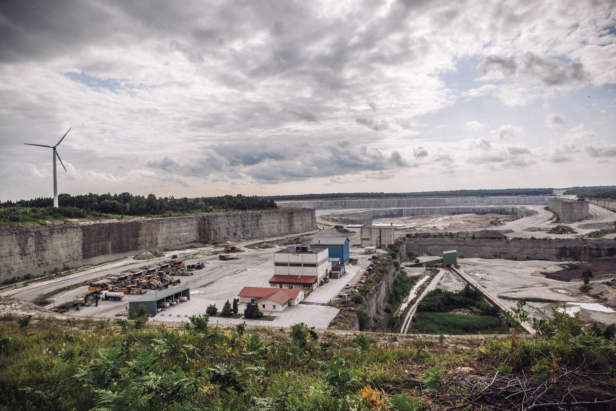 A large pale quarry with small industrial buildings and a wind turbine beneath clouds.