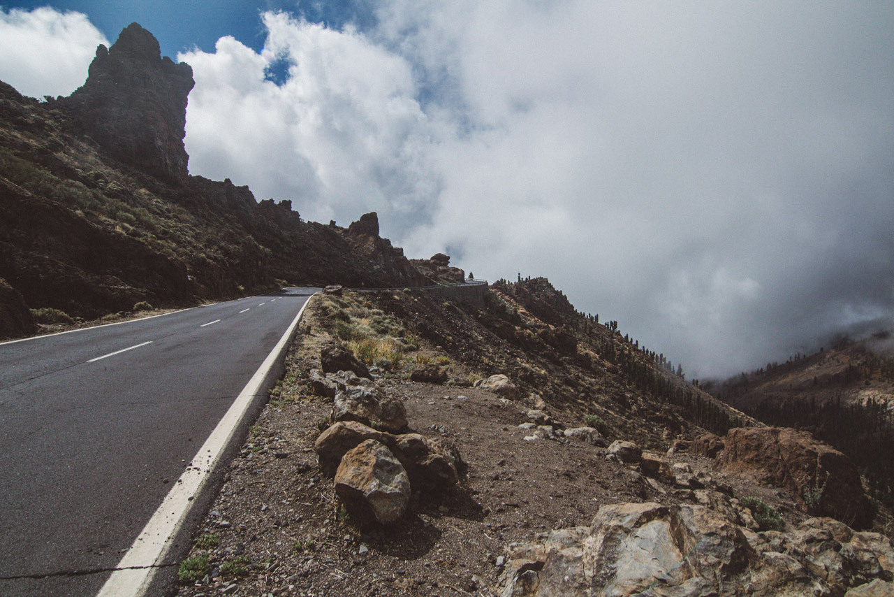 A mountain road follows a rocky edge beside large white clouds.